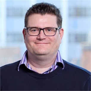 James Wood in glasses, a navy jumper and purple striped shirt, in front of a window